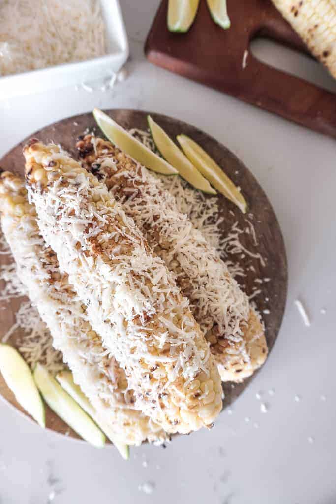 Mexican Street Corn pictured on a wooden plate with sliced limes. The Mexican Street corn is covered in chili powder and thinly grated cotija cheese