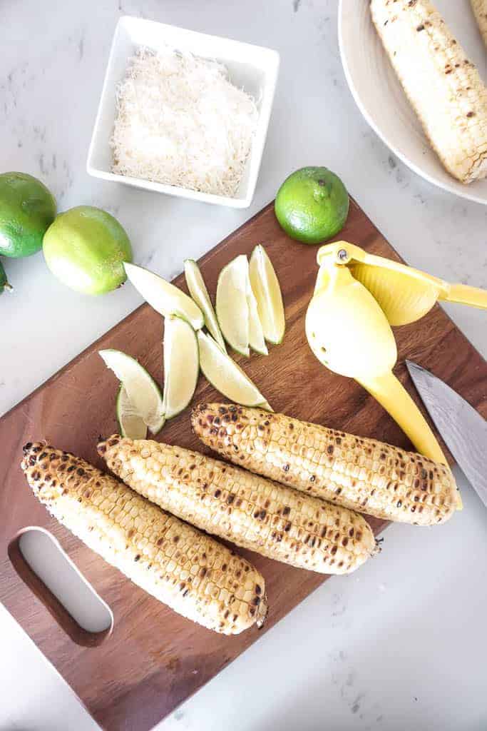 Ingredients for Mexican street corn laid out on the counter. On a wood cutting board, there are sliced limes and corn and off to the side there are full limes and mayonnaise