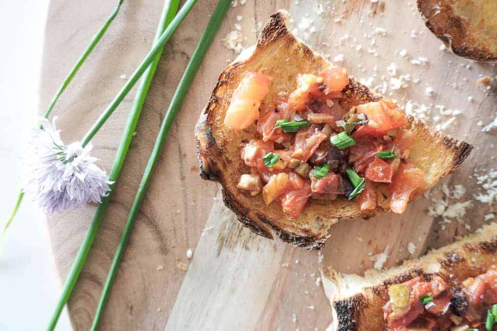 Signed Samantha's olive and bruschetta recipe - a birdseye view of olive bruschetta spread on a toasted baguette. There are two pieces placed on a cutting board with olives in a bowl sitting next to it.