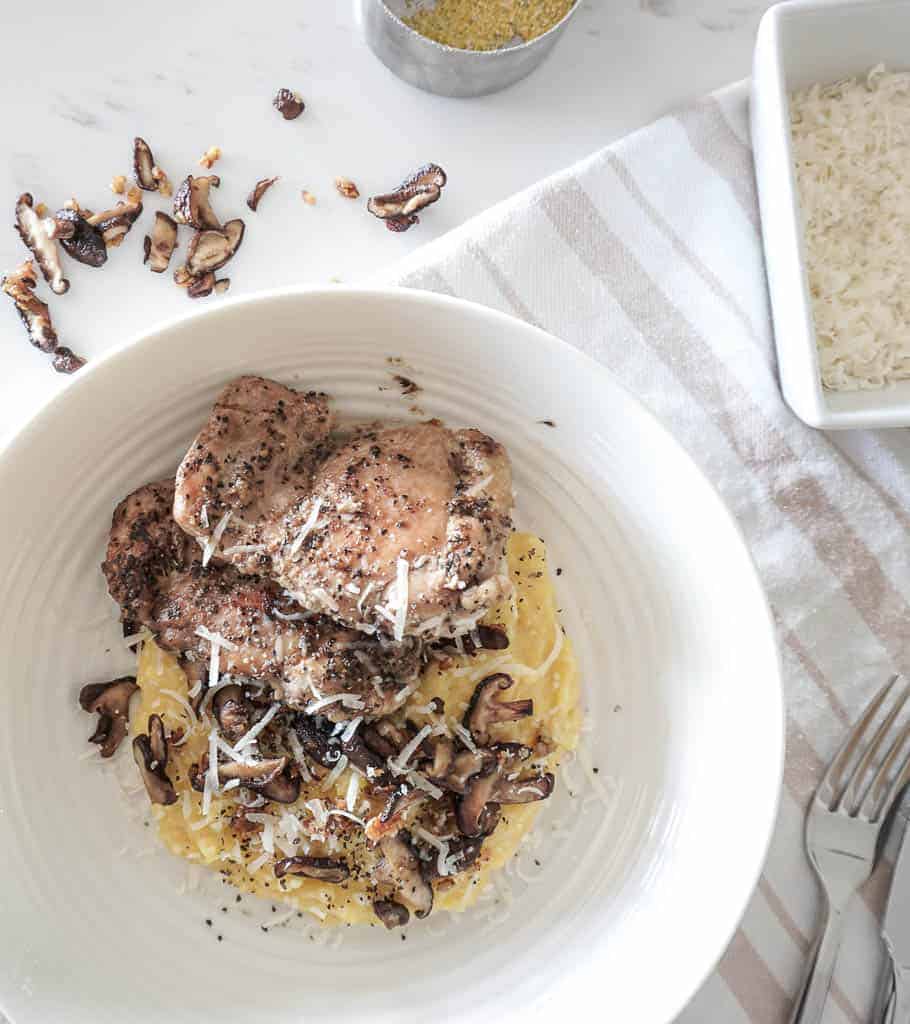 Signed Samantha's chicken and polenta recipe - an overhead shot of polenta in a low pasta bowl with mushrooms, parmesan and topped with chicken with mushrooms on the counter in the background as well as a bowl of parmesan cheese.