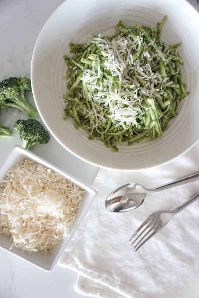 Signed Samantha kale pasta recipe plated and ready to serve. In the image there is a bowl full of parmesan, broccoli, and a low bowl full of green pasta sauce and spaghetti noodles.