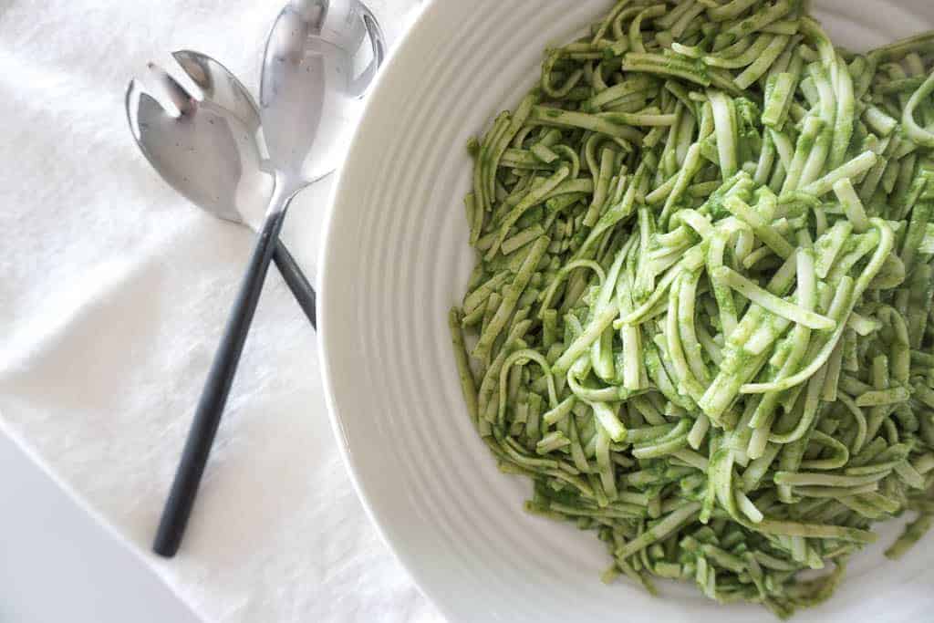 Kale pasta recipe from Signed Samantha that's all ready to serve. The green spaghetti noodles are in a round serving bowl with two serving utensils off to the side