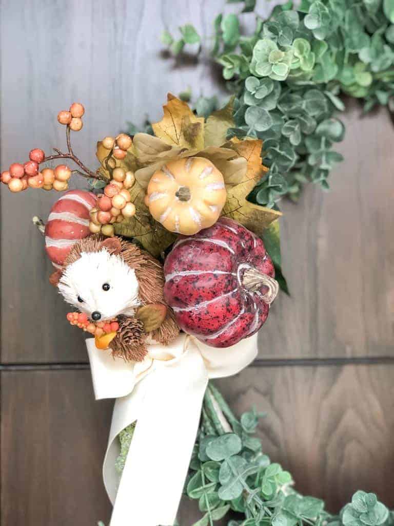 Signed Samantha's instructions on how to make a wreath step by step are included and limited items are needed. Pictured is 1/6th of the wreath with green eucalyptus, and another faux long green plant. The long green plant is tied in a bow with a cream coloured ribbon. Right above the ribbon are a bunch of mini pumpkins, red, orange, and yellow, and a faux hedgehog.