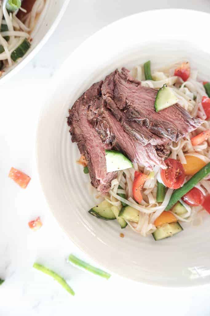 Signed Samantha's Rice Noodle Salad recipe picture which includes rice noodles, chopped cucumber, green beans, red peppers, and flank steak pictured in a white pasta bowl. Off to the side is the serving bowl of the noodles.