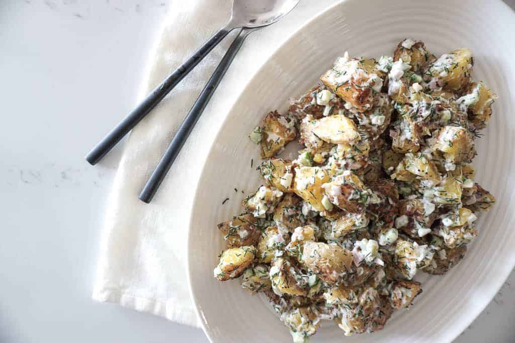 Vegan potato salad by Signed Samantha. A flatlay image of potato salad in an oval bowl, sitting on top of a cream linen napkin with two serving spoons next to it.