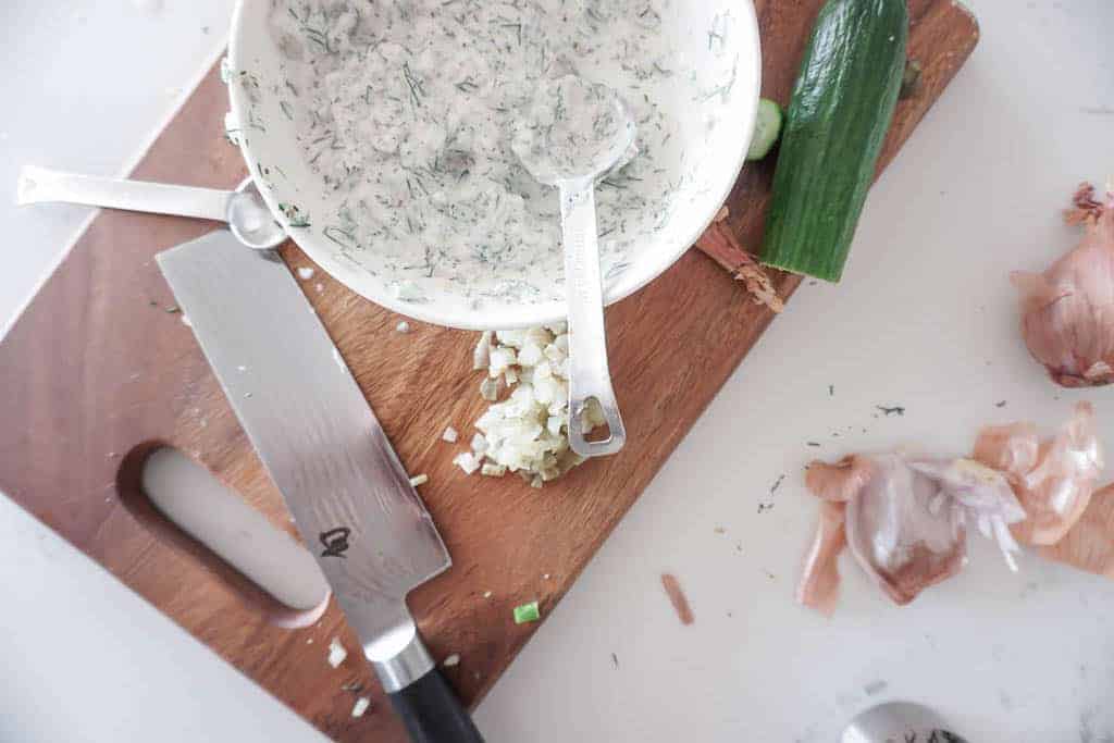 Signed Samantha's creamy dill sauce for her vegan potato salad. An overhead shot of the creamy dill mixture, with shallot skins, cucumber and a knife in the background.