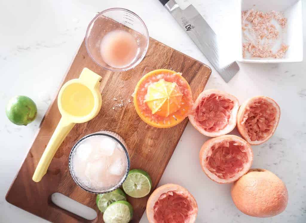 Signed Samantha's easy paloma recipe in the making. On the counter, there are four halves of grapefruits whose juices have been squeezed out, one whole grapefruit, a knife, and a bowl with rimmer in it. There is a cutting board and on the cutting board are three lime halves whose juice has been squeezed, two hand juicers, and a cup filled with the paloma.