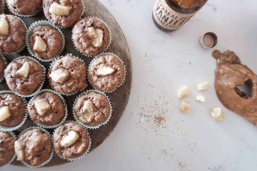 Overhead shot of Signed Samantha's gluten free banana muffins. The mini muffins are pictured on a circular platter, with 1/4 sliced bananas on top, 1/4 sliced bananas in the side, a spoon with batter laid on the counter, and a bottle of vanilla pictured.