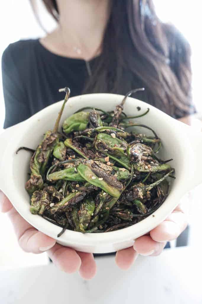 Signed Samantha's blistered shishito peppers are pictured in a white bowl being held by two hands facing the camera.