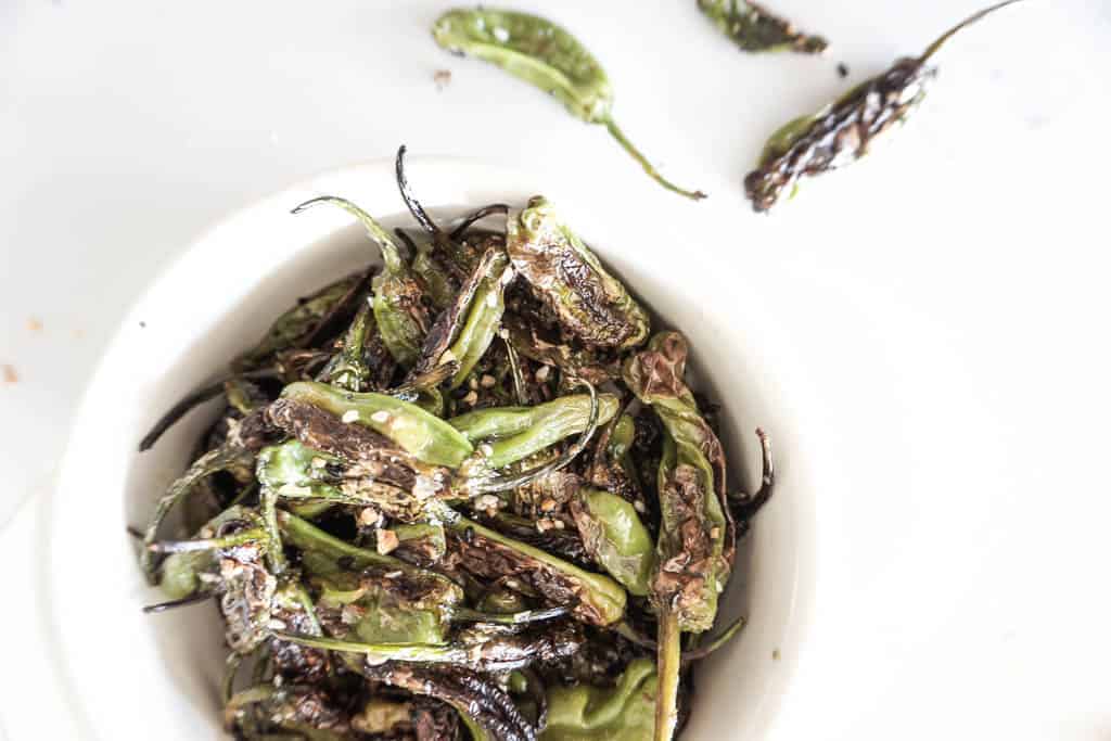 Signed Samantha's blistered shishito peppers are pictured in a white bowl with shishito peppers in the background on the counter.