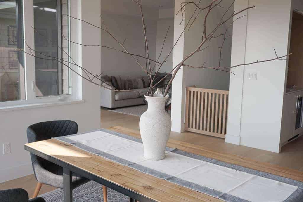 Signed Samantha's DIY stone vase is pictured on top of her dining room table which is a wood table with a runner on it. There are many large branches coming of the stone vase. In the background is Samantha's living room.