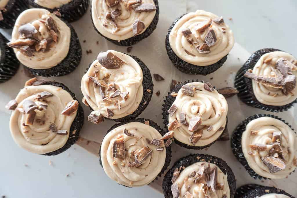 Signed Samantha's gluten-free chocolate cupcakes pictured on a wood cutting board. The chocolate cupcakes have a peanutbutter mascarpone frosting on them with crushed up skor bar on top.