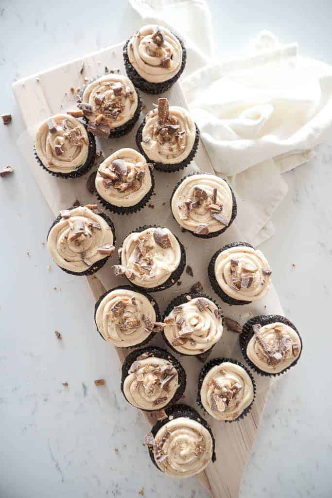 Signed Samantha's Gluten-Free Chocolate Cupcakes pictured on a wood cutting board. The chocolate cupcakes have a peanutbutter mascarpone frosting on them with crushed up skor bar on top.