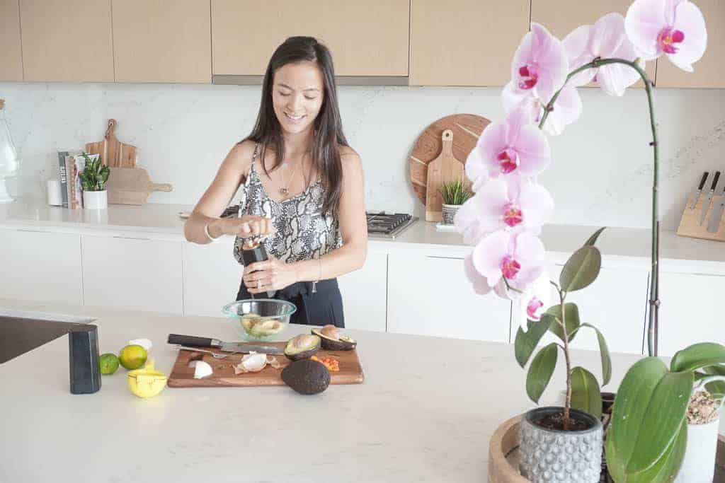 Signed Samantha standing in her kitchen making gaucamole. She is grinding pepper in a bowl with avocados. In the background are her kitchen counters - the subject of the post is how to style kitchen counterotps