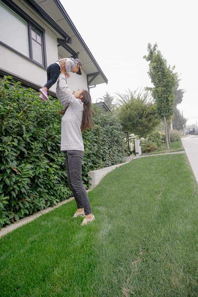 Signed Samantha is gently throwing her daughter Sloane in the air while she stands on the grass, next to a large hedge. Samantha is discussing what I've learned about motherhood