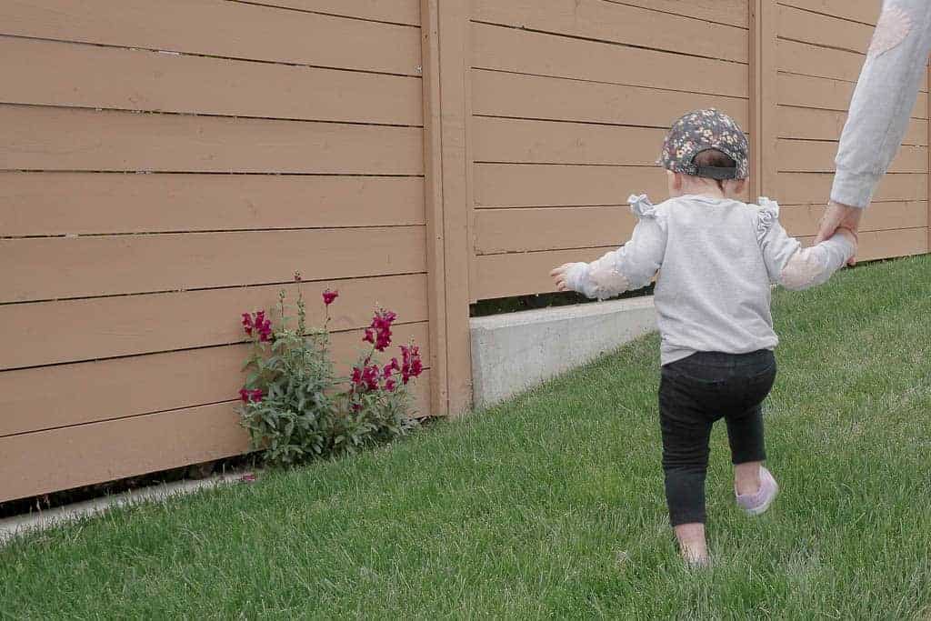 Signed Samantha's daughter Sloane is exploring, about to go smell the red flowers that are growing out from behind the brown fence. Samantha is discussing what I've learned about motherhood.