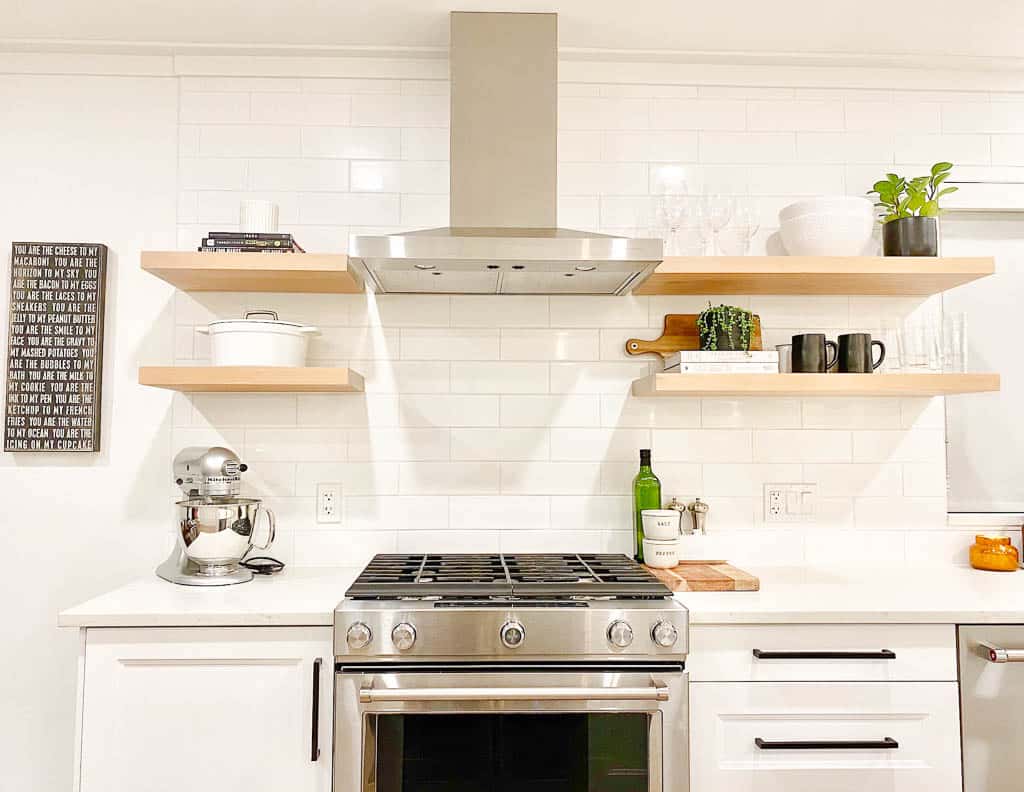 Signed Samantha's friend Caitlin's kitchen countertop style pictured is the stove and on one side there is a kitchen aid mixer, and the other side has olive oil salt and pepper and butter. Above both sides are open floating shelves which has a pot on one shelf, cook books, plants, and candles on the other.