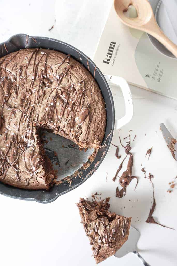 Signed Samantha's Skillet Hazelnut Brownie Blondie Bars pictured in a white cast iron skillet, and a piece cut out sitting on the counter. In the background there is a box of parchment paper that says kana on it.