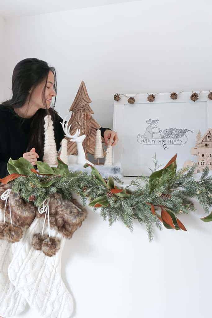 Signed Samantha placing a D.I.Y mini christmas tree onto her mantle. There are five beige trees pictured made of yarn, a white reindeer, a wooden tree, and a picture of a moose. On the edge of the mantle is garland, and stockings.