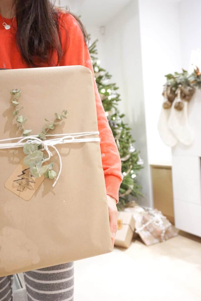 Signed Samantha holding up a holiday gift showing her D.I.Y holiday gift tags. The gift is wrapped in craft paper with dried greens.