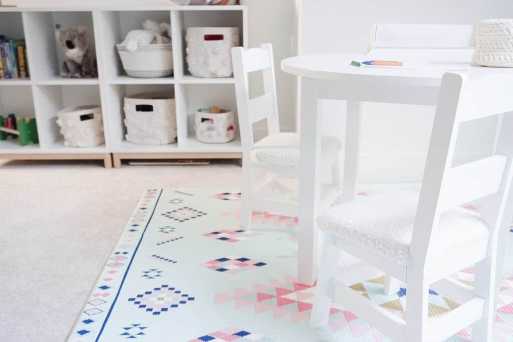 D.I.Y Kids Chair Cushion is pictured on two white chairs next to a round kids table on top of a play mat. In the background is a shelving unit with kids toys on it.