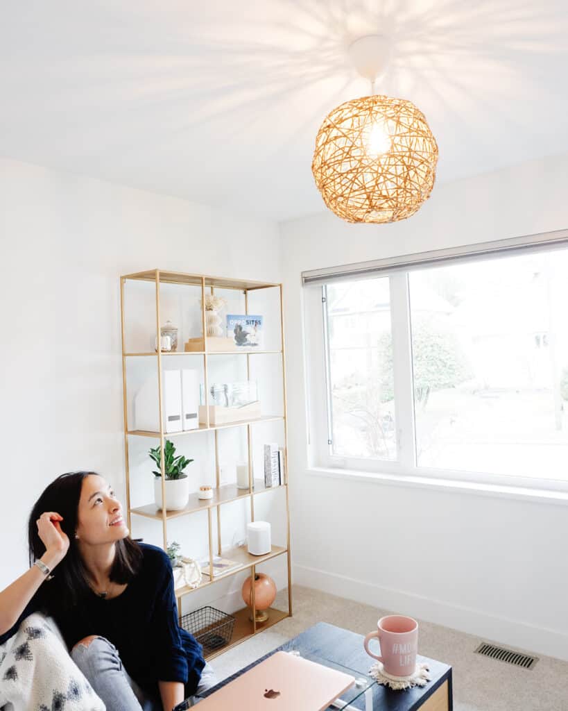 Signed Samantha standing below her D.I.Y pendant light in her home office which has a desk, laptop, and shelf in the background.