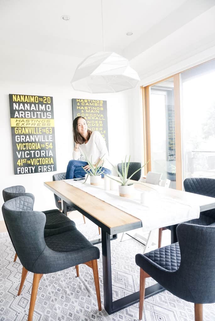 Signed Samantha's dining room tour - a view of her dining room with a carpet, white pendant light, plants, and bus rolls - Samantha is sitting on the dining room table