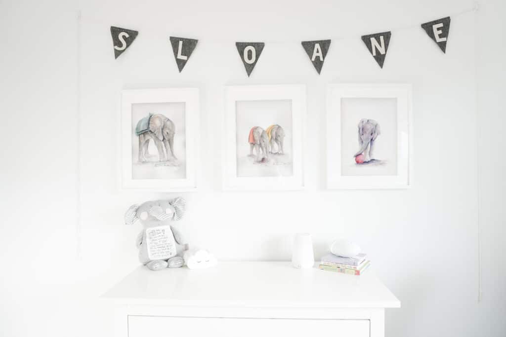 Sloane's nursery tour, pictured is a beautiful banner with Sloane's name on it, three water colour elephants hanging on the walls, and a white dresser with an elephant, sound machine, and diffisuer