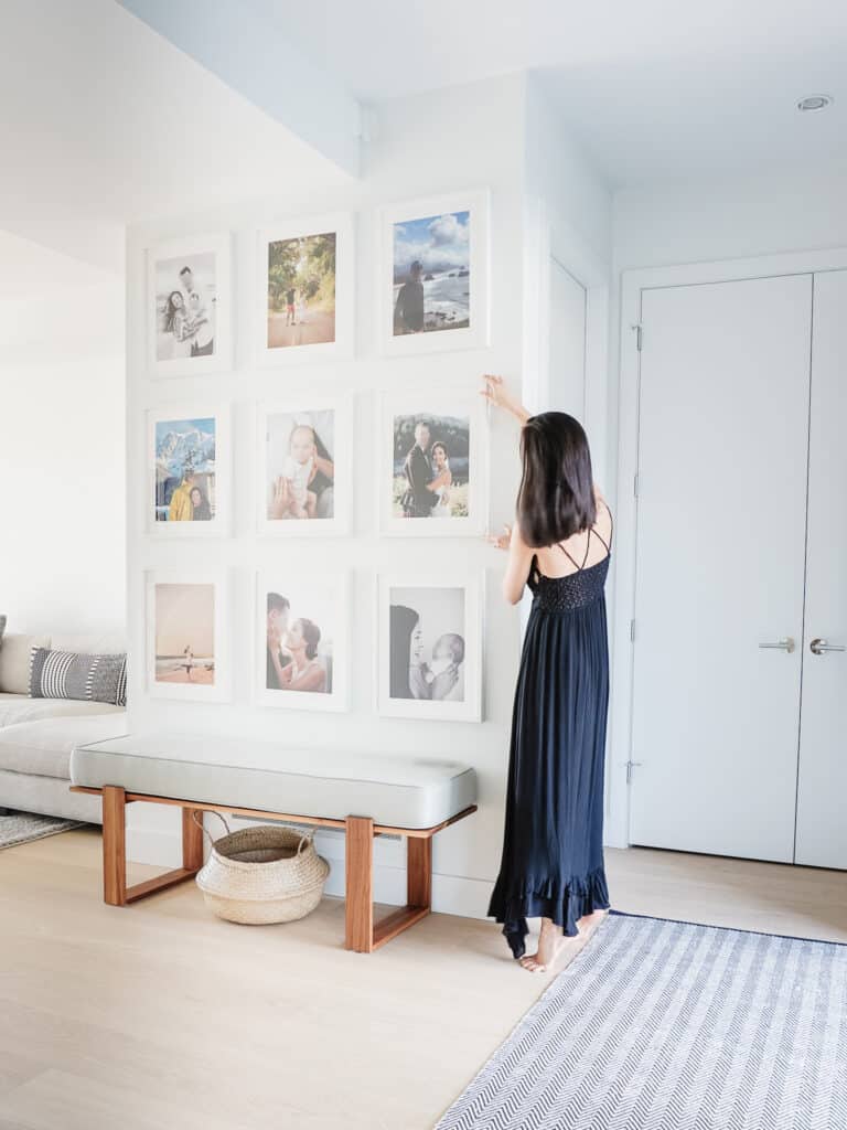 Signed Samantha adjusting her gallery wall of nine white picture frames, each matted, containing family photostalking about how to make your house feel like a home