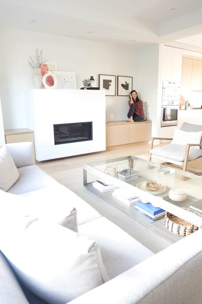 Signed Samantha gives a living room tour - here is she sharing a full shot of her living room including a sofa, coffee table, two chairs, and end tables. Everything is neutral in colour. Samantha Potter is sitting next to the fireplace on a built in bench.