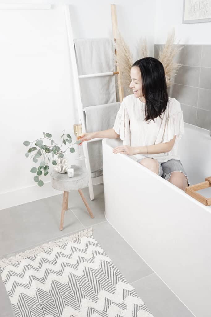 Siged Samantha Potter sitting in a bath tub next to her beautiful DIY concrete table that she made. It is about 3 inches thick with three wooden legs. There is also a white vase with eucaylptus, a champagne flute, and candle on there. In the background is a ladder with towels on it, and in the foregroud there is a chevron rug.