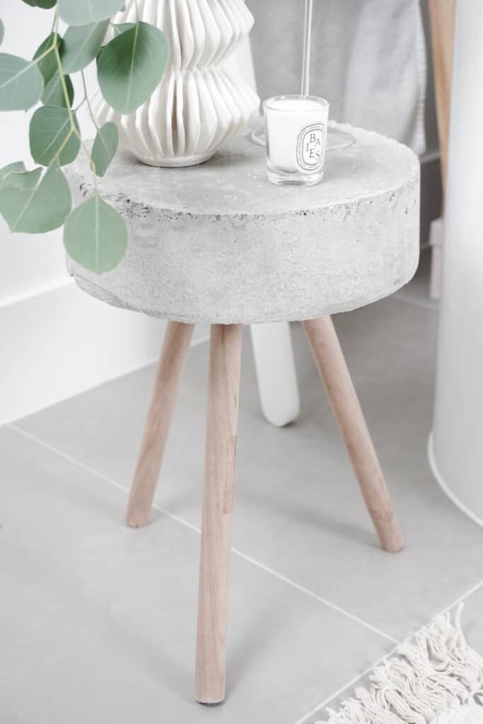 A beautiful concrete side table that Signed Samantha Potter made. It is about 3 inches thick with three wooden legs. There is also a white vase with eucaylptus, a champagne flute, and candle on there.