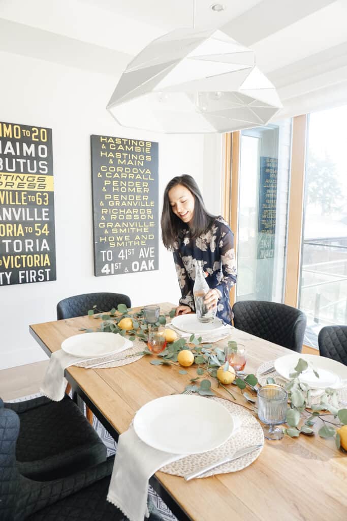 Signed Samantha sharing some home decor updates for spring including how to update your tablescape for this season. Pictured is her wood table with eucaylptus and lemons as well as neutral napkins and placemats, white plates and coloured glassware. Samantha is standing in the background pouring water.