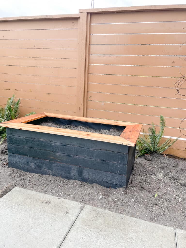 Garden box sitting outside in the garden bed. It is a black garden box with a non-painted cedar cap. Approx. 4 ft. long x 2 ft deep.