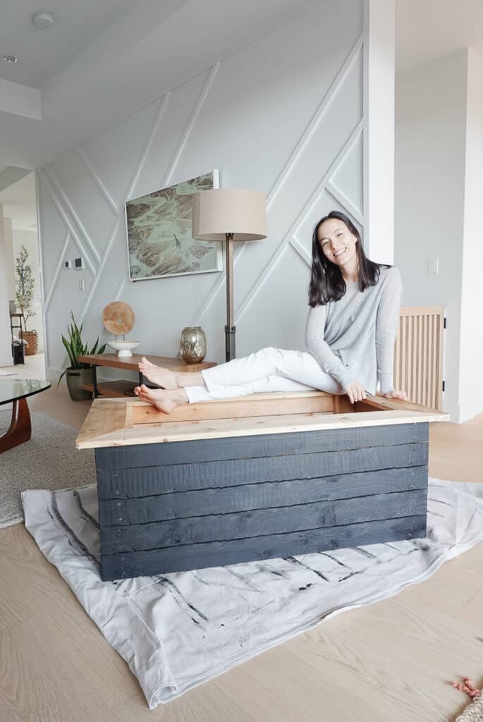 Signed samantha sitting on her garden box in her house in front of a white wall with a geometric pattern made from white moulding. The garden box is in her house for this picture because she doesn't like the colour of her fence.