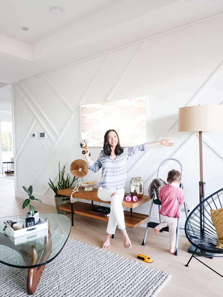 Signed Samantha teaching you how to install board and batten while sitting on her media console in front of her modern board and batten that is going in different directions on the wall. Her toddler daughter is climinb a ladder next to her playing with tools