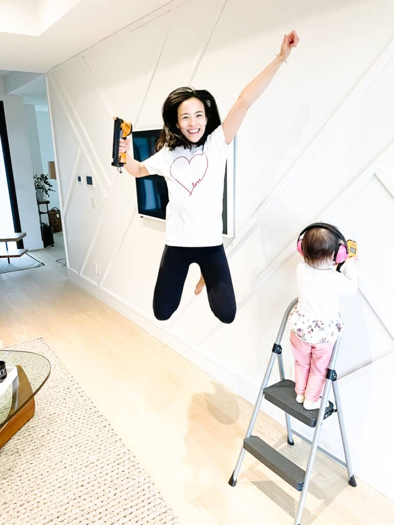 Signed Samantha teaches you how to install board and batten she is jumping for joy with her nail gun (not connected and emptied for safety reasons) in front of her completed wall with her daughter standing in front of it.