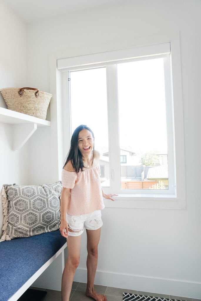 Sharing Samantha Potter's DIY Window Trim around the window in her mudroom. It is unpainted and not filled and sanded, yet. Next to a bench with a blue cushion on it and a decorative pillow. Samantha is standing in front of the window smiling.