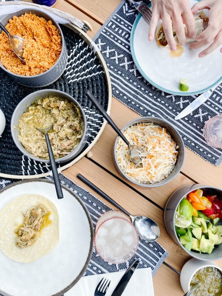 Slow Cooker Chicken Verde in a bowl served on a table with rice, taco shells, avocado and tomatoes, and cheese. Each in a bowl. Hands are in the picture making a taco.