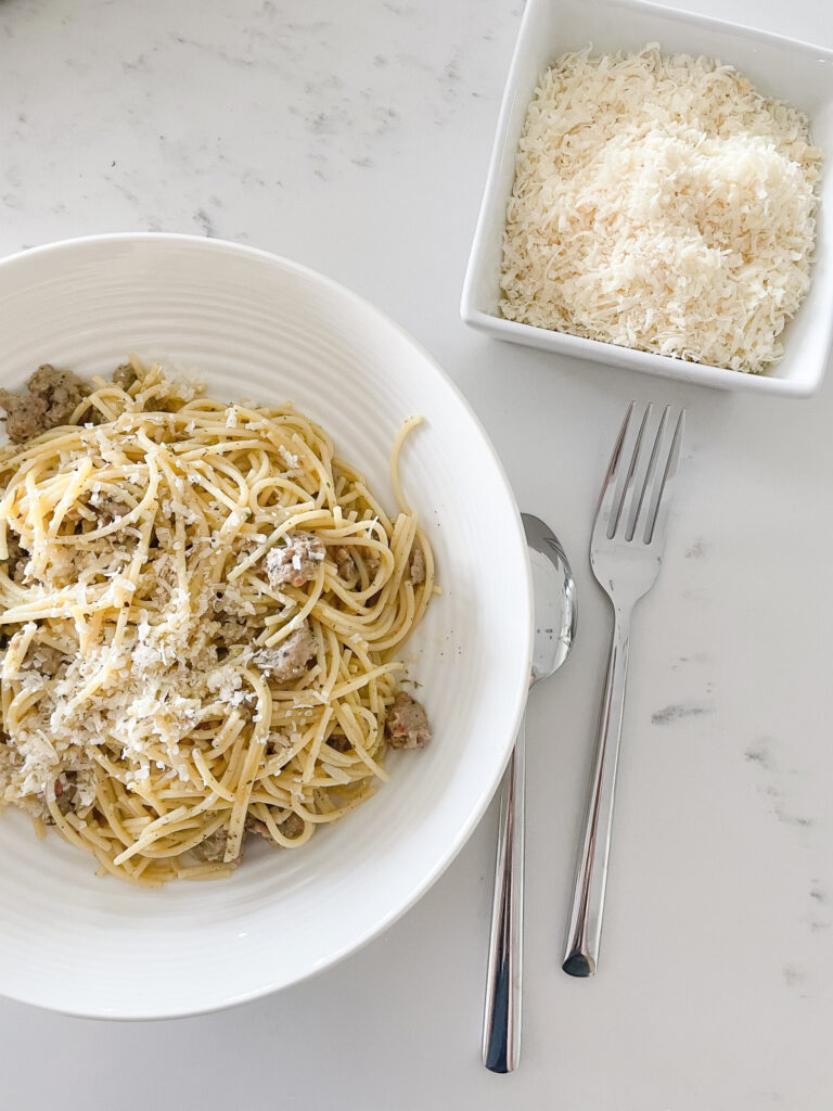 Sausage carbonara in a low pasta bowl with a fork and a spoon next to it as well as a bowl of parmesan cheese