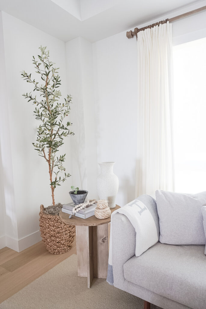 Alward side table from studio mcgee.. excep its a DIY version. It is a pine wood table with an "x" lengthwise for the legs and a round top. It is decorated with a vase, two books, a plant and a set of wooden beads. An olive tree is in the back ground. and a grey sofa next to it.