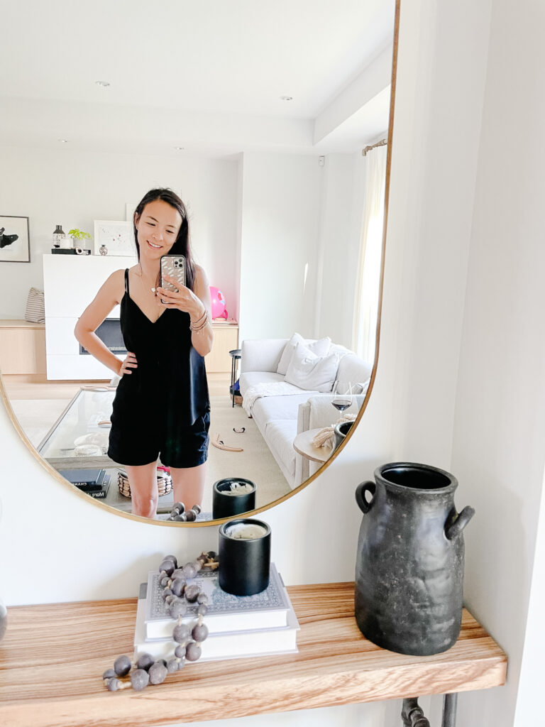 Signed Samantha standing in front of her diy antique vase and mirror of her living room where there is a mess behind her.