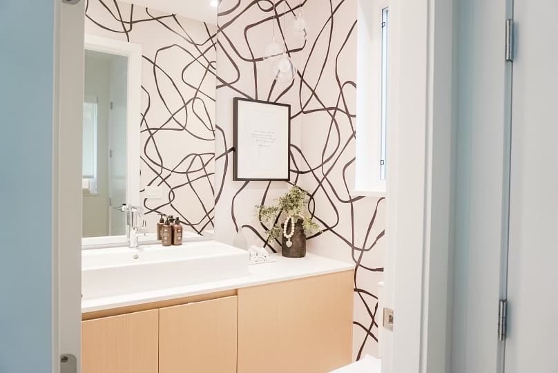 Peel and stick wallpaper in an abstract pattern on the wall - the background is a pinky taupe and the patterns are black. There is a black picture frame on the wall, a vase on the counter in the bathroom, soap, and towels.