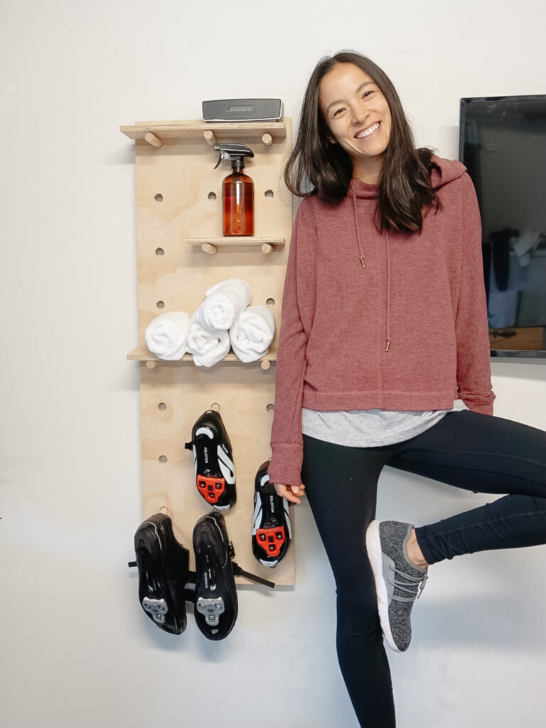 Signed Samantha standing in front of her DIY pegbaord with her drill in hand. There is a spray bottle and towels on her pegboard