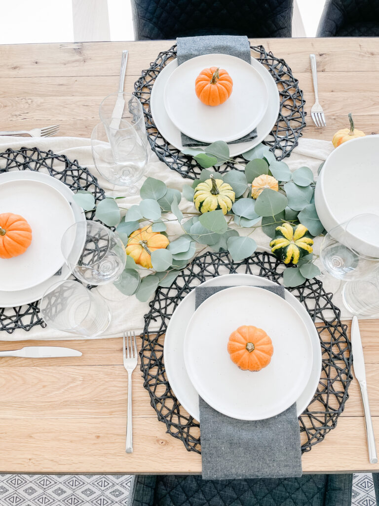 Setting the table for thanksgiving in Signed Samantha's house includes a runner down the middle of the table, black placemats, white dishwear, pumpkins, eucyalptus, and wine galsses