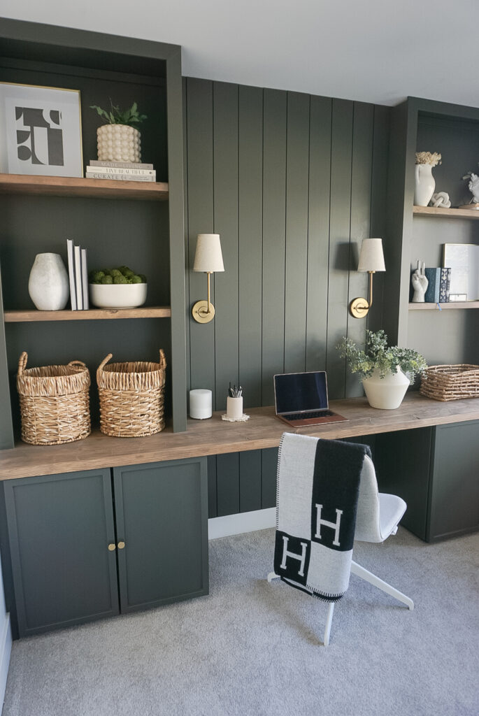 built in home office set up in a deep mossy green with shiplap and lighting at the back, bookshelves on either side and neutral decor throughout.