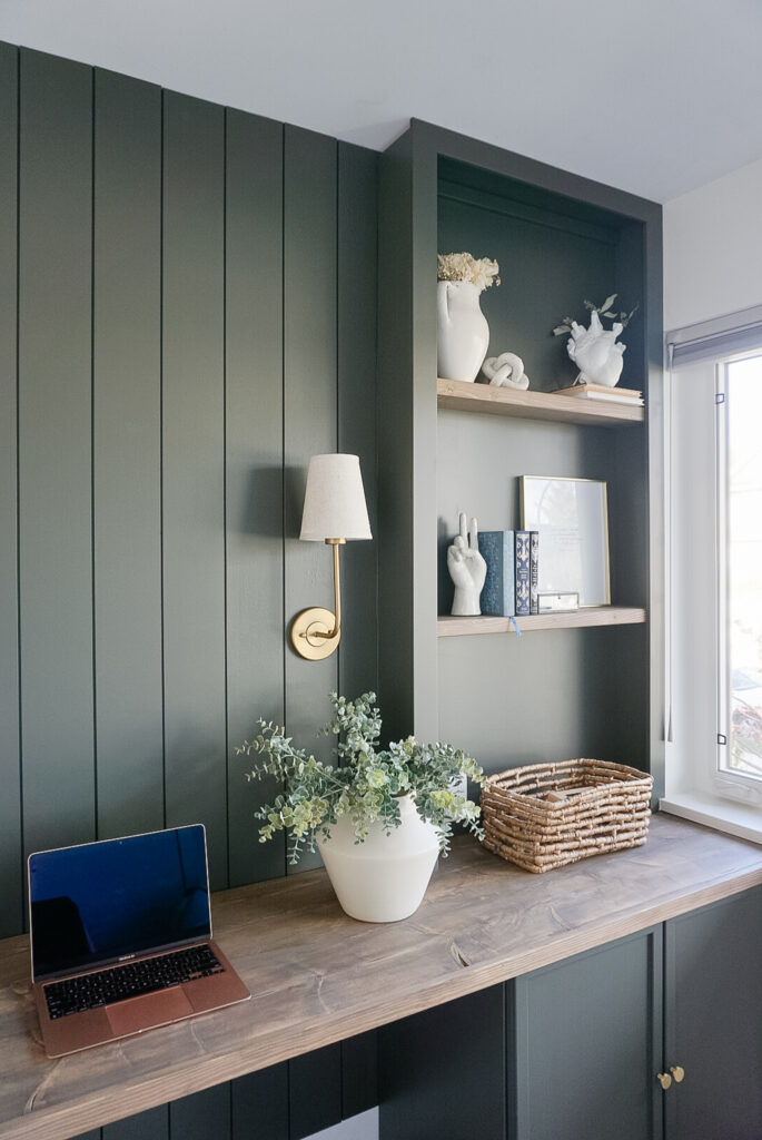 built in home office set up in a deep mossy green with shiplap and lighting at the back, bookshelves on either side and neutral decor throughout.