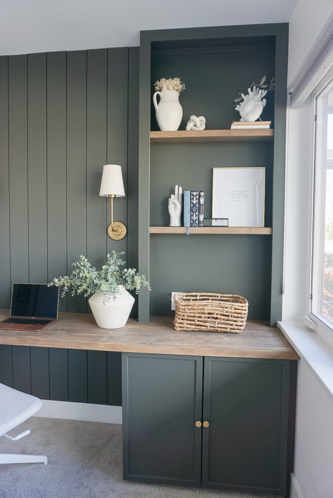 built in home office set up in a deep mossy green with shiplap and lighting at the back, bookshelves on either side and neutral decor throughout.