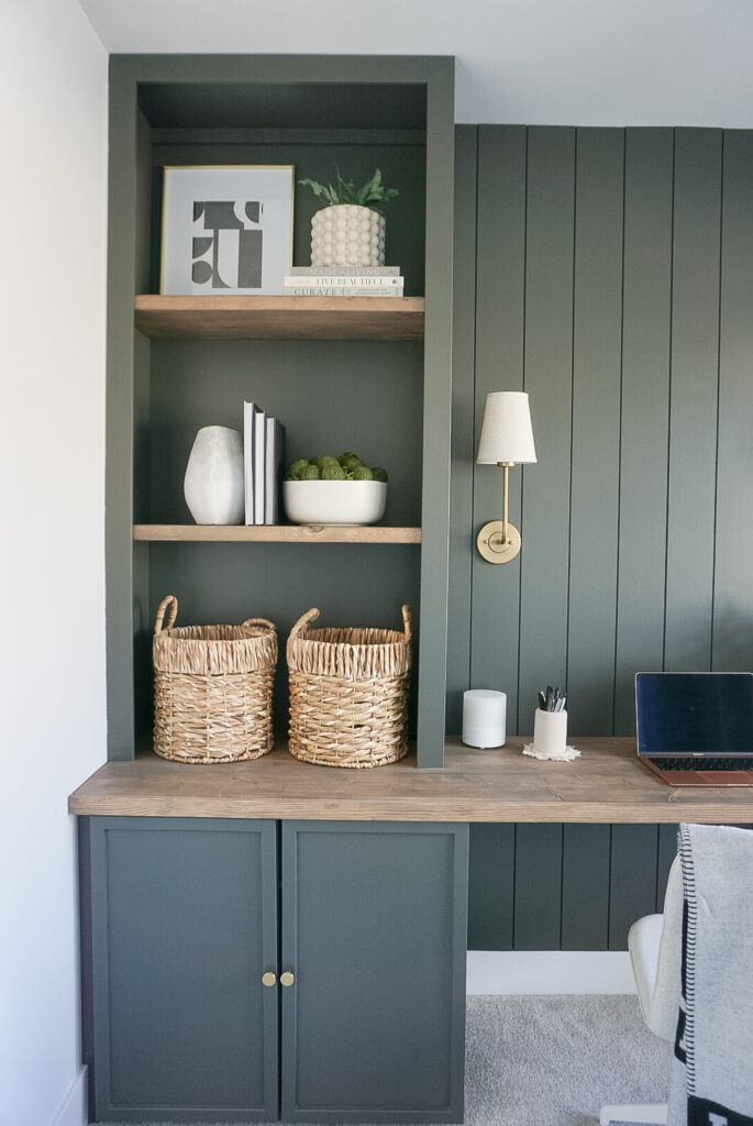 DIY Built-In Bookshelf in a green colour with shelves stained in a darker wood tone. The built-in bookshelf is sitting on top of a wood desktop. Decorated with baskets on the lowest shelf, bowls, books, plants, and pictures above.