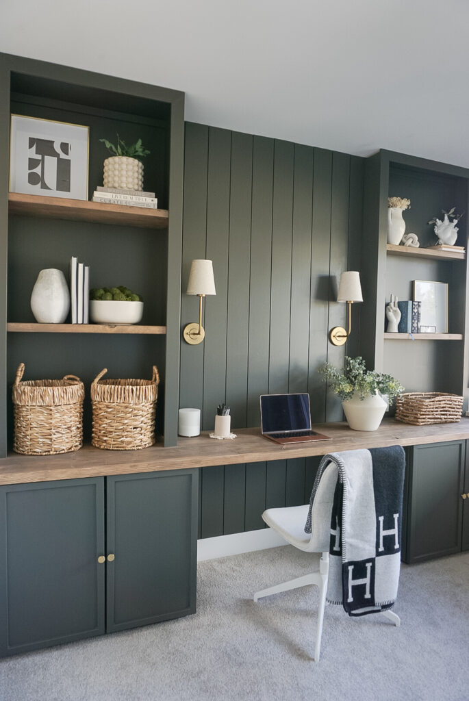 built in home office set up in a deep mossy green with shiplap and lighting at the back, bookshelves on either side and neutral decor throughout.